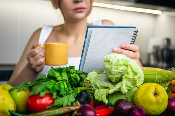 A person writes in a mindfulness journal next to a plate of fruit. A kitchen with healthy groceries fills the background, promoting a mindful and healthy relationship with food.