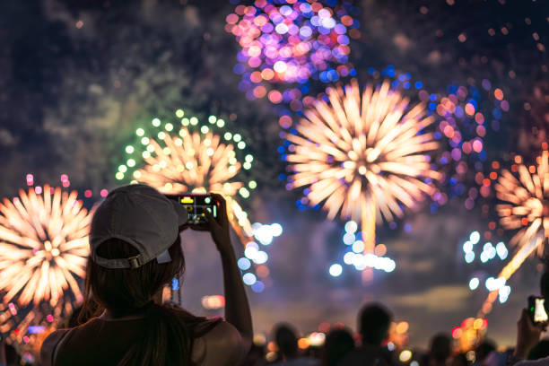 A person gazing upwards at a night sky illuminated by a dazzling display of fireworks. In their hand, they hold a spent sparklers, its embers fading